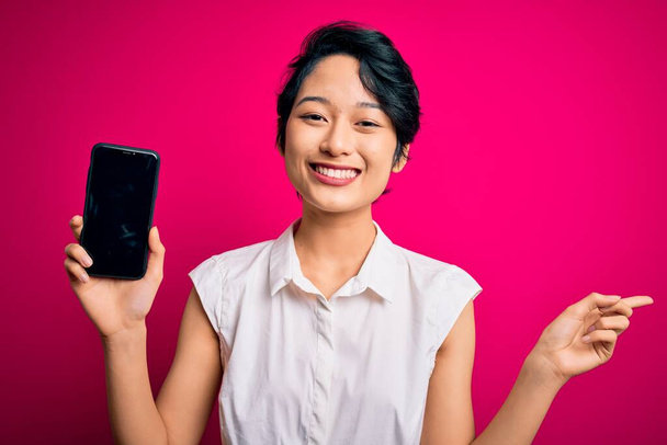 Jovem bela menina asiática segurando smartphone mostrando tela sobre fundo rosa isolado muito feliz apontando com a mão e o dedo para o lado
 - Foto, Imagem