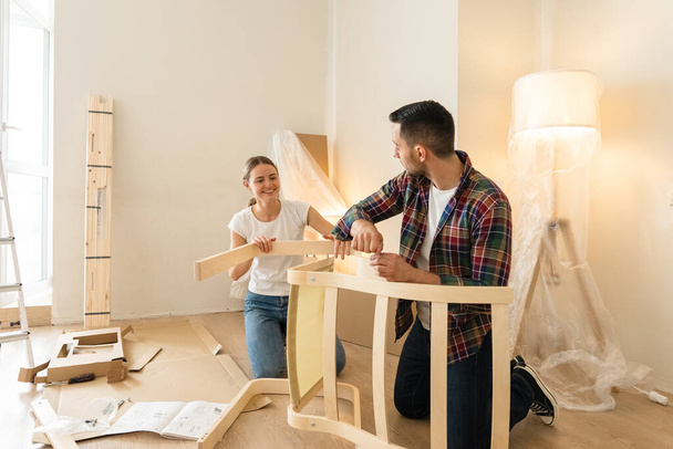Happy Smiling Couple Collects Furniture as a Team. Girl Helps to Assemble Chair Details. Moving to New Apartment, Young Family Assemble Furniture - 写真・画像