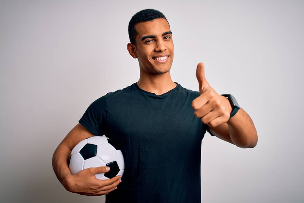 Handsome african american man playing footbal holding soccer ball over white background approving doing positive gesture with hand, thumbs up smiling and happy for success. Winner gesture. - Photo, Image