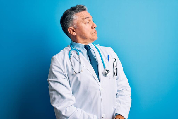 Middle age handsome grey-haired doctor man wearing coat and blue stethoscope looking to the side with arms crossed convinced and confident - Foto, imagen