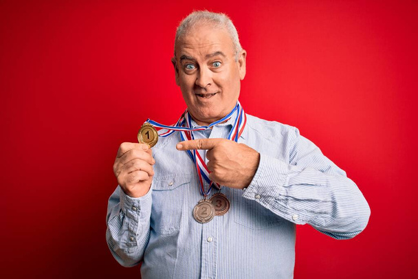 meia idade hoary campeão homem vestindo medalhas de pé sobre isolado fundo vermelho muito feliz apontando com a mão e o dedo
 - Foto, Imagem