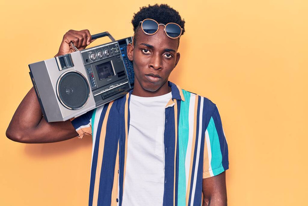 Młody, afrykański mężczyzna trzymający boombox, słuchający muzyki myślącej i trzeźwego wyrazu twarzy wyglądający na pewnego siebie  - Zdjęcie, obraz