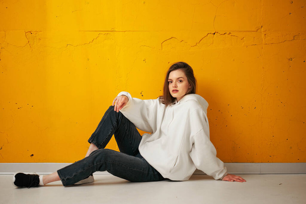 Young pretty calm relaxing girl in a white hoodie poses sitting on the floor against a yellow concrete wall. - Fotografie, Obrázek