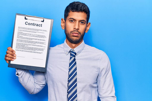 Young latin man holding clipboard with contract document thinking attitude and sober expression looking self confident  - Foto, Imagem