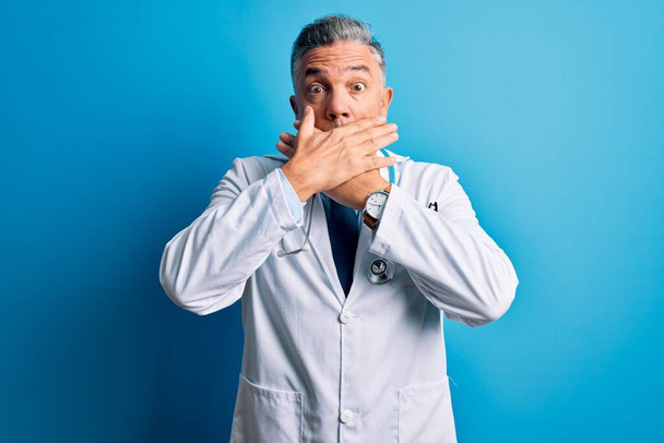 Keski-ikä komea harmaa tukka lääkäri mies yllään takki ja sininen stetoskooppi järkyttynyt peittää suun kädet erehdyksen. Salainen käsite. - Valokuva, kuva