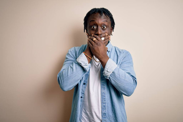 Mladý pohledný africký Američan v neformální džínové košili na bílém pozadí šokované zakrývající ústa s rukama za omyl. Tajný koncept. - Fotografie, Obrázek