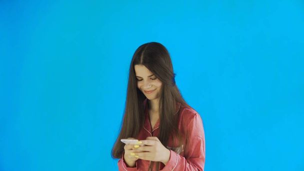 Jeune femme heureuse en pyjama utilise smartphone sur fond bleu - Photo, image