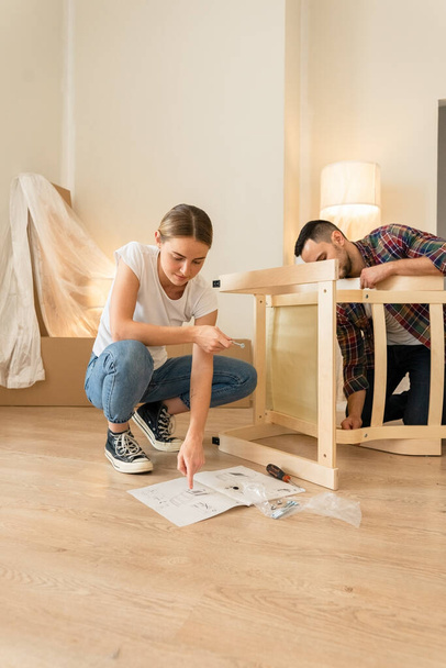 Happy Smiling Couple Collects Furniture as a Team. Girl Helps to Assemble Chair Details. Moving to New Apartment, Young Family Assemble Furniture - Фото, изображение