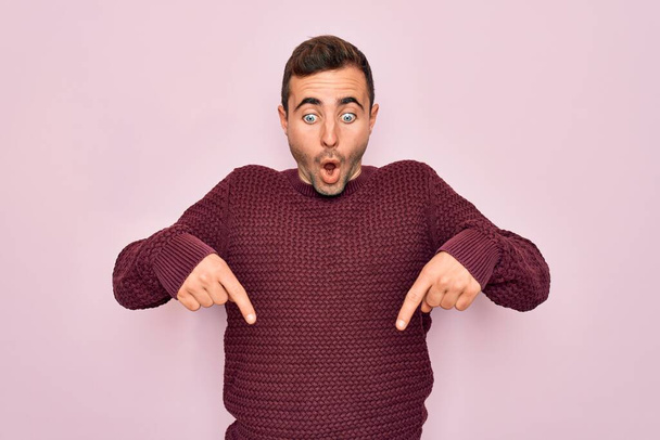 Jonge knappe man met blauwe ogen met casual trui over roze achtergrond naar beneden wijzend met vingers met reclame, verrast gezicht en open mond - Foto, afbeelding