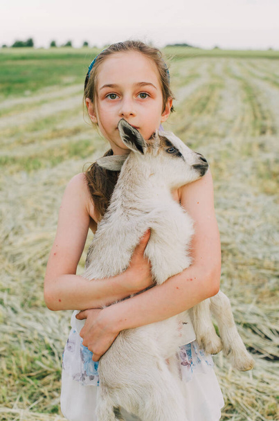 girl with baby goat on farm outdoors. Love and care. Village animals. happy child hugs goat, concept of unity of nature and man. Friendship of child and animals. Happy childhood. Girl and goat - 写真・画像