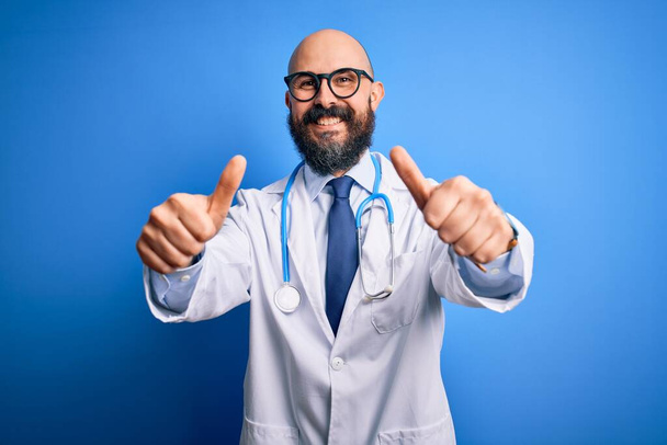 Knappe kale arts met baard die een bril draagt en stethoscoop over een blauwe achtergrond die positief gebaar met de hand goedkeurt, glimlacht en blij is voor succes. Winnaar gebaar. - Foto, afbeelding