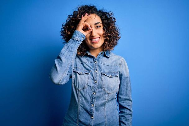 Молодая красивая кудрявая арабская женщина в джинсовой рубашке, стоящей на синем фоне, делает хорошо жест с улыбкой на руке, глаза смотрят сквозь пальцы с счастливым лицом
. - Фото, изображение