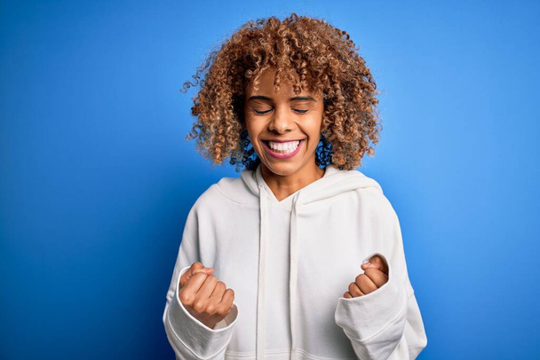 Gyönyörű afro-amerikai sportos nő visel alkalmi pulóver felett kék háttér izgatott a siker karját, és csukott szemmel ünnepli győzelem mosolyogva. Nyertes koncepció. - Fotó, kép