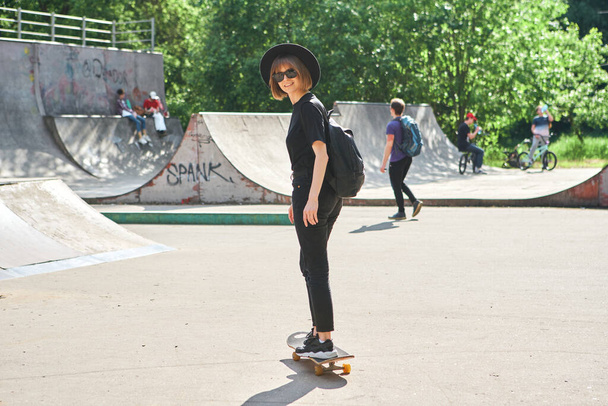 Pretty smiling teenage girl in hat and sunglasses with backpack rides skateboard in skatepark active and sport lifestyle - Φωτογραφία, εικόνα