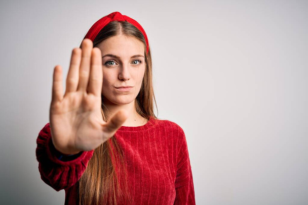 Молодая красивая рыжая женщина в красном свитере и диадеме на жёлтом фоне делает остановку петь с ладонью руки. Предупреждающее выражение с негативным и серьезным жестом на лице
. - Фото, изображение
