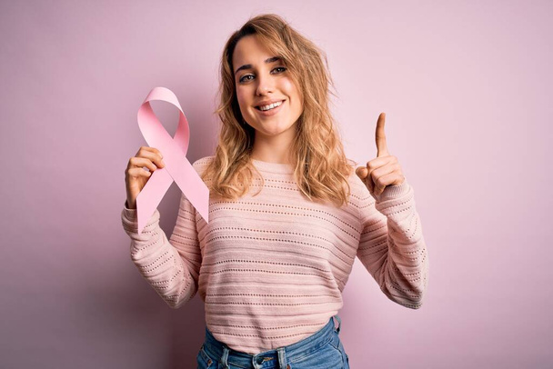 Junge schöne blonde Frau mit rosa Krebsband Symbol über isoliertem Hintergrund überrascht mit einer Idee oder Frage Zeigefinger mit glücklichem Gesicht, Nummer eins - Foto, Bild