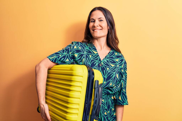 Young beautiful brunette tourist woman holding airplane cabin suitcase over yellow background looking positive and happy standing and smiling with a confident smile showing teeth - Fotó, kép