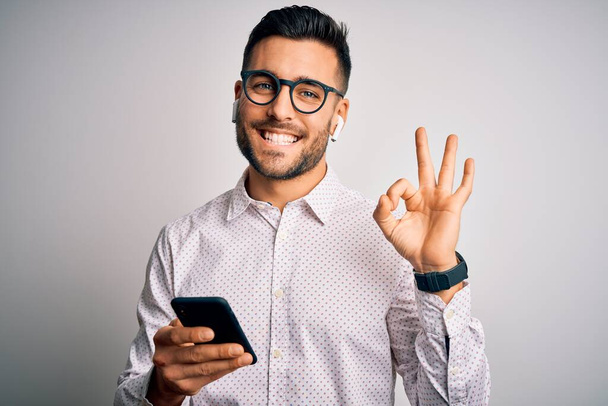 Mladý pohledný muž s brýlemi při konverzaci pomocí smartphonu a sluchátek dělá v pořádku znamení s prsty, vynikající symbol - Fotografie, Obrázek