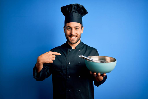 Joven cocinero con barba vistiendo uniforme usando batidor y tazón sobre fondo azul con la cara sorpresa apuntando con el dedo hacia sí mismo
 - Foto, imagen