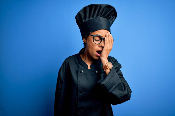 Joven chef afroamericana mujer con uniforme de cocina y sombrero sobre fondo azul Bostezo cansado cubriendo media cara, ojo y boca con la mano. La cara duele de dolor
. - Foto, imagen