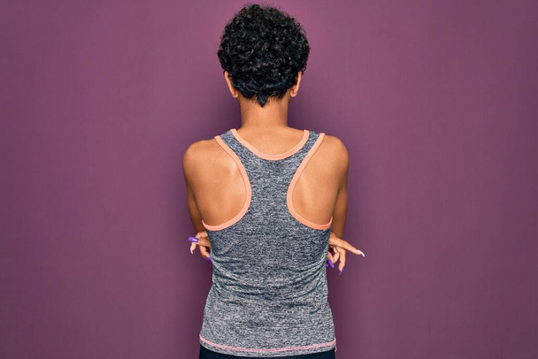 Jeune belle afro afro-américaine sportive faisant de l'exercice portant des vêtements de sport debout vers l'arrière regardant loin avec les bras croisés - Photo, image