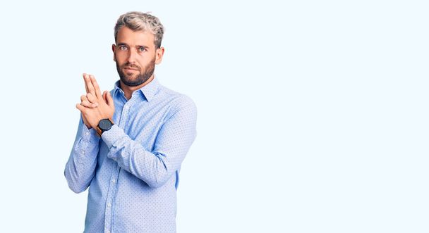 Young handsome blond man wearing elegant shirt holding symbolic gun with hand gesture, playing killing shooting weapons, angry face  - Photo, Image