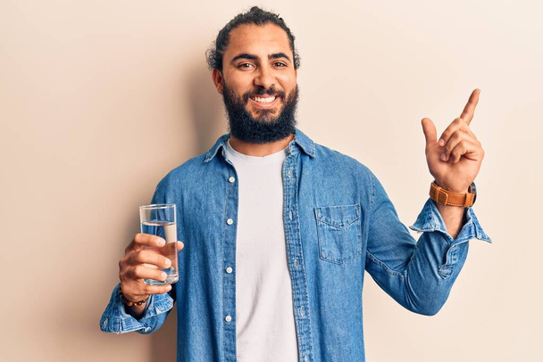Giovane arabo bere bicchiere d'acqua sorridente felice indicando con mano e dito di lato  - Foto, immagini