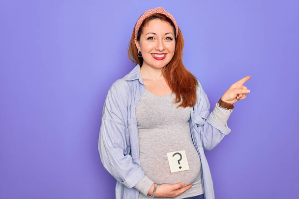 Jeune belle rousse femme enceinte attend bébé avec point d'interrogation sur le ventre très heureux pointant avec la main et le doigt sur le côté - Photo, image