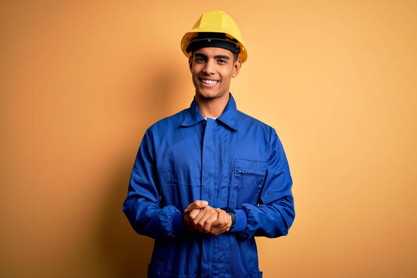 Joven trabajador afroamericano guapo vestido con uniforme azul y casco de seguridad con las manos juntas y los dedos cruzados sonriendo relajado y alegre. Éxito y optimismo
 - Foto, Imagen