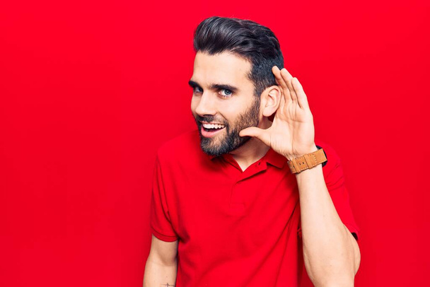 Young handsome man with beard wearing casual polo smiling with hand over ear listening and hearing to rumor or gossip. deafness concept.  - Photo, Image