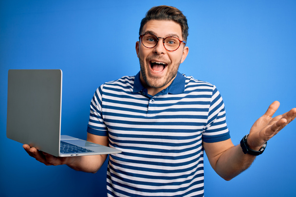 コンピューターのノートパソコンで働く青い目をした青い目の若いビジネスマンはとても幸せで興奮し、大きな笑顔で叫んで勝利を祝う勝者の表現と手を上げ - 写真・画像