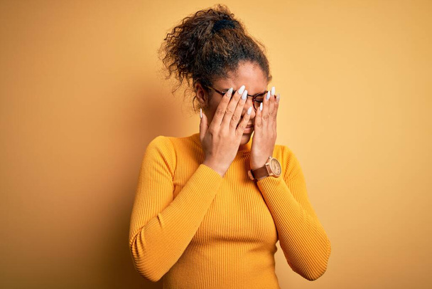 Young beautiful african american girl wearing sweater and glasses over yellow background rubbing eyes for fatigue and headache, sleepy and tired expression. Vision problem - Photo, Image