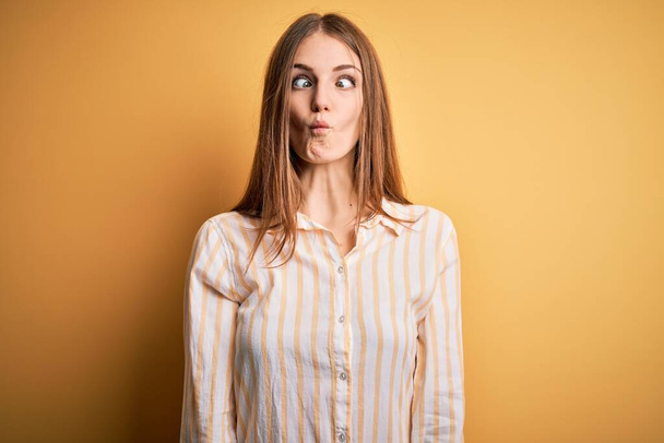 Young beautiful redhead woman wearing casual striped shirt over isolated yellow background making fish face with lips, crazy and comical gesture. Funny expression. - Foto, immagini