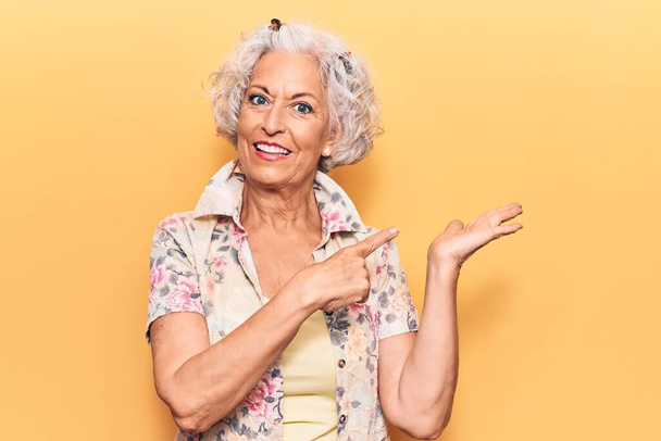 Старша сіро-волохата жінка в повсякденному одязі здивована і посміхається камері, представляючи рукою і вказуючи пальцем
.  - Фото, зображення