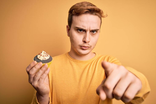 Молодой красивый рыжий мужчина ест сладкий шоколадный десерт на желтом фоне указывая пальцем в камеру и для вас, уверенный жест выглядит серьезным
 - Фото, изображение