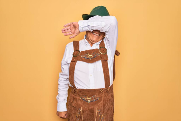Senior grey-haired man wearing german traditional octoberfest suit over yellow background covering eyes with arm smiling cheerful and funny. Blind concept. - Foto, Imagen