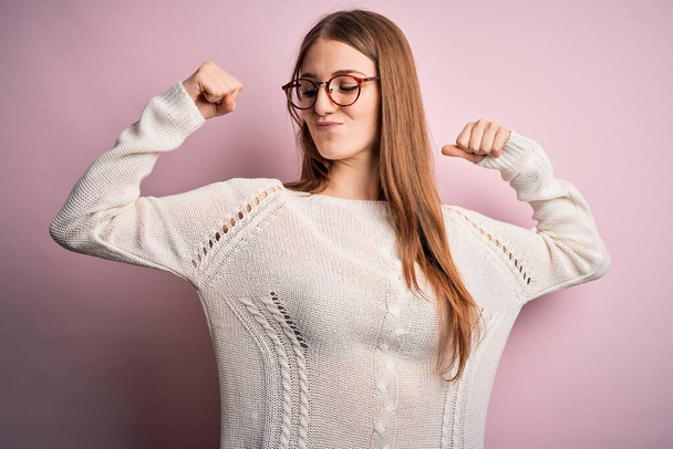 Mujer pelirroja hermosa joven con suéter casual y gafas sobre fondo rosa mostrando los músculos de los brazos sonriendo orgulloso. Concepto Fitness
. - Foto, imagen