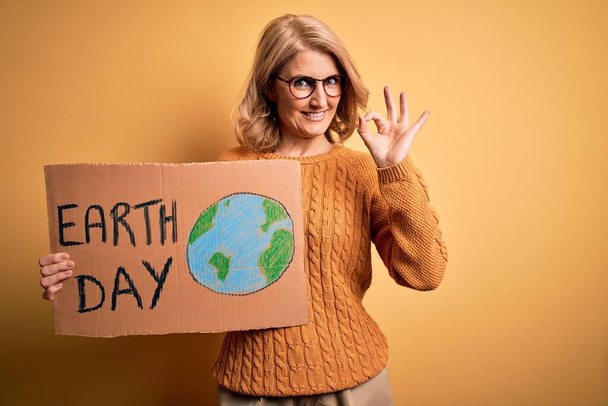 Mulher loira de meia idade pedindo o ambiente segurando banner com mensagem do dia da terra fazendo sinal ok com os dedos, excelente símbolo
 - Foto, Imagem