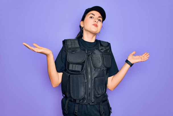 Jeune femme de la police portant gilet pare-balles de sécurité uniforme sur fond violet expression vide et confus avec les bras et les mains levées. Concept de doute. - Photo, image