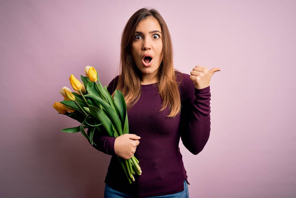 Young blonde woman holding romantic bouquet of yellow tulips flowers over pink background Surprised pointing with hand finger to the side, open mouth amazed expression. - Photo, Image