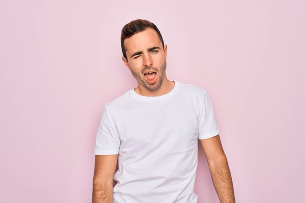 Όμορφος άνδρας με μπλε μάτια φορώντας casual λευκό t-shirt στέκεται πάνω από ροζ φόντο κλείνει το μάτι κοιτάζοντας την κάμερα με σέξι έκφραση, χαρούμενο και χαρούμενο πρόσωπο. - Φωτογραφία, εικόνα