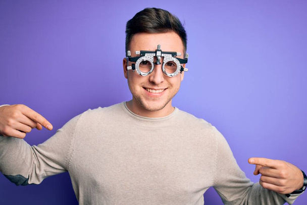 Jeune homme caucasien beau portant des lunettes optométriques sur fond violet regardant confiant avec le sourire sur le visage, se montrant du doigt fier et heureux. - Photo, image