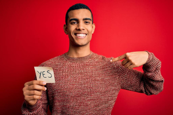 Young handsome african american man holding reminder paper with yes message with surprise face pointing finger to himself - Foto, imagen