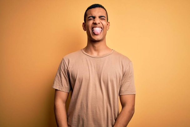 Jovem homem afro-americano bonito vestindo t-shirt casual em pé sobre fundo amarelo colando a língua para fora feliz com expressão engraçada. Conceito de emoção
. - Foto, Imagem