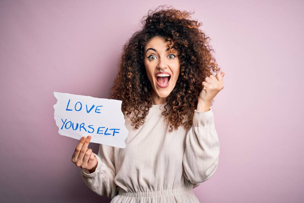 Young beautiful woman with curly hair and piercing holding paper with love yourself message screaming proud and celebrating victory and success very excited, cheering emotion - Photo, Image