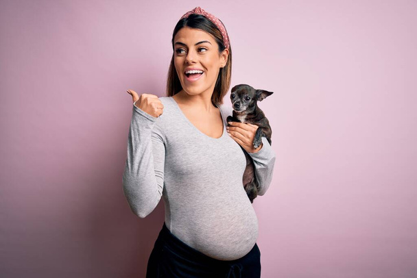 Joven hermosa morena embarazada esperando bebé sosteniendo chihuahua cachorro señalando y mostrando con el pulgar hacia el lado con la cara feliz sonriendo
 - Foto, imagen