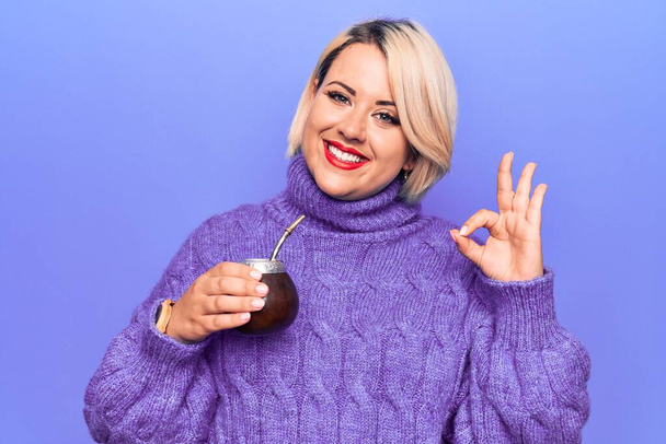 Beautiful blonde plus size woman drinking mate infusion beverage over purple background doing ok sign with fingers, smiling friendly gesturing excellent symbol - Photo, Image