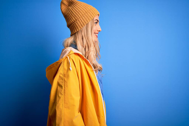 Young beautiful blonde woman wearing raincoat for rainy weather over blue background looking to side, relax profile pose with natural face with confident smile. - 写真・画像