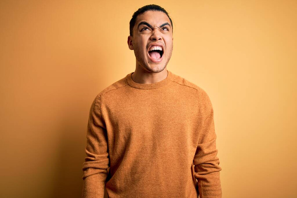 Молодий бразільський чоловік у звичайному светрі стоїть над жовтим фоном, злий і божевільний, кричачи розстроєним і розлюченим, кричачи від гніву. Жорстокість і агресивна концепція. - Фото, зображення