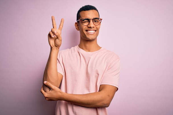 Komea afrikkalainen amerikkalainen mies päällään rento t-paita ja lasit vaaleanpunainen tausta hymyillen onnellinen kasvot silmää kameralle tekemässä voiton merkki sormin. Numero kaksi.. - Valokuva, kuva